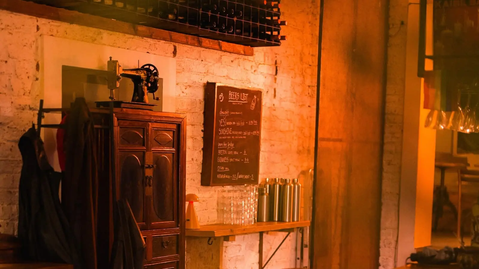 The shop floor at night: whitewashed brick, a chalkboard beer list, a dresser with an old sewing machine on top and stools along the bar